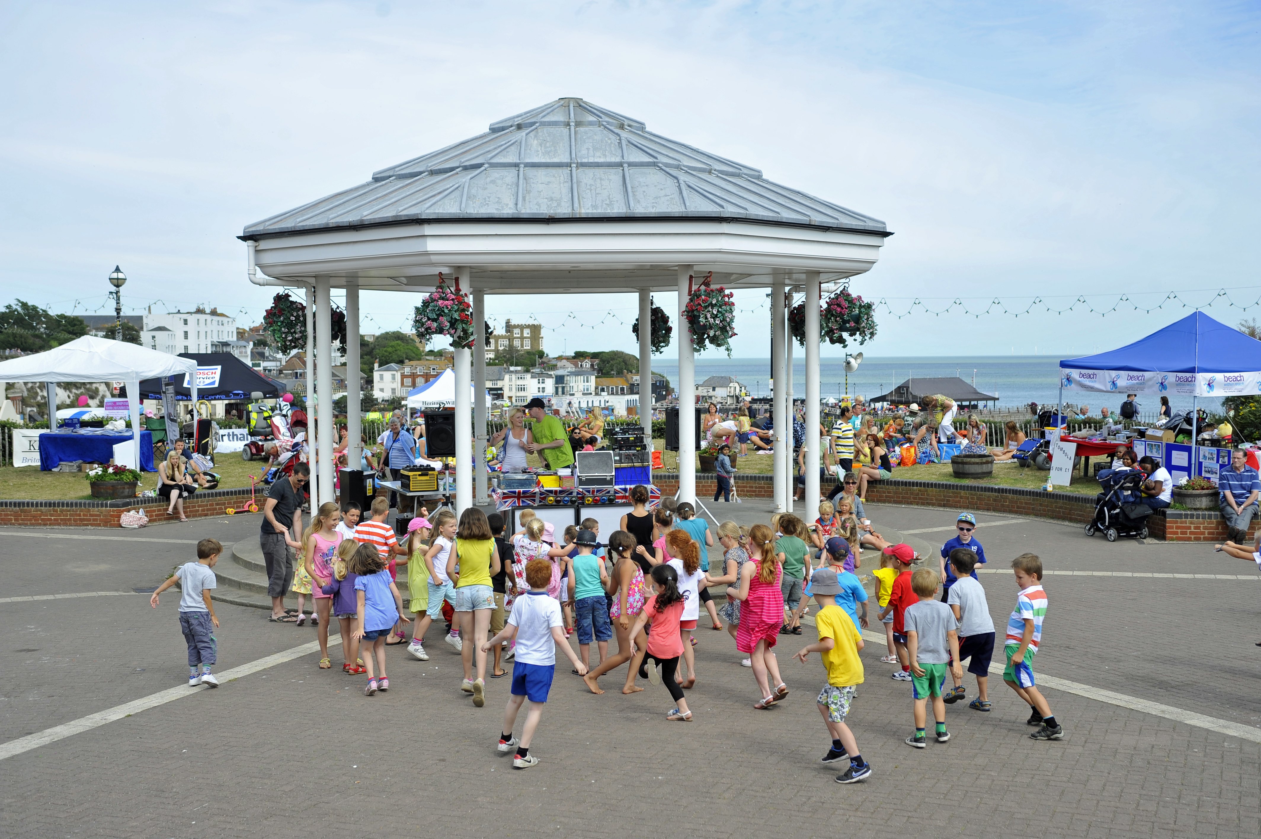 Children dancing in front of white bandstand with dj playing music. Variety of gazebos/stalls either side of bandstand selling/promoting things.  Light blue/grey sky