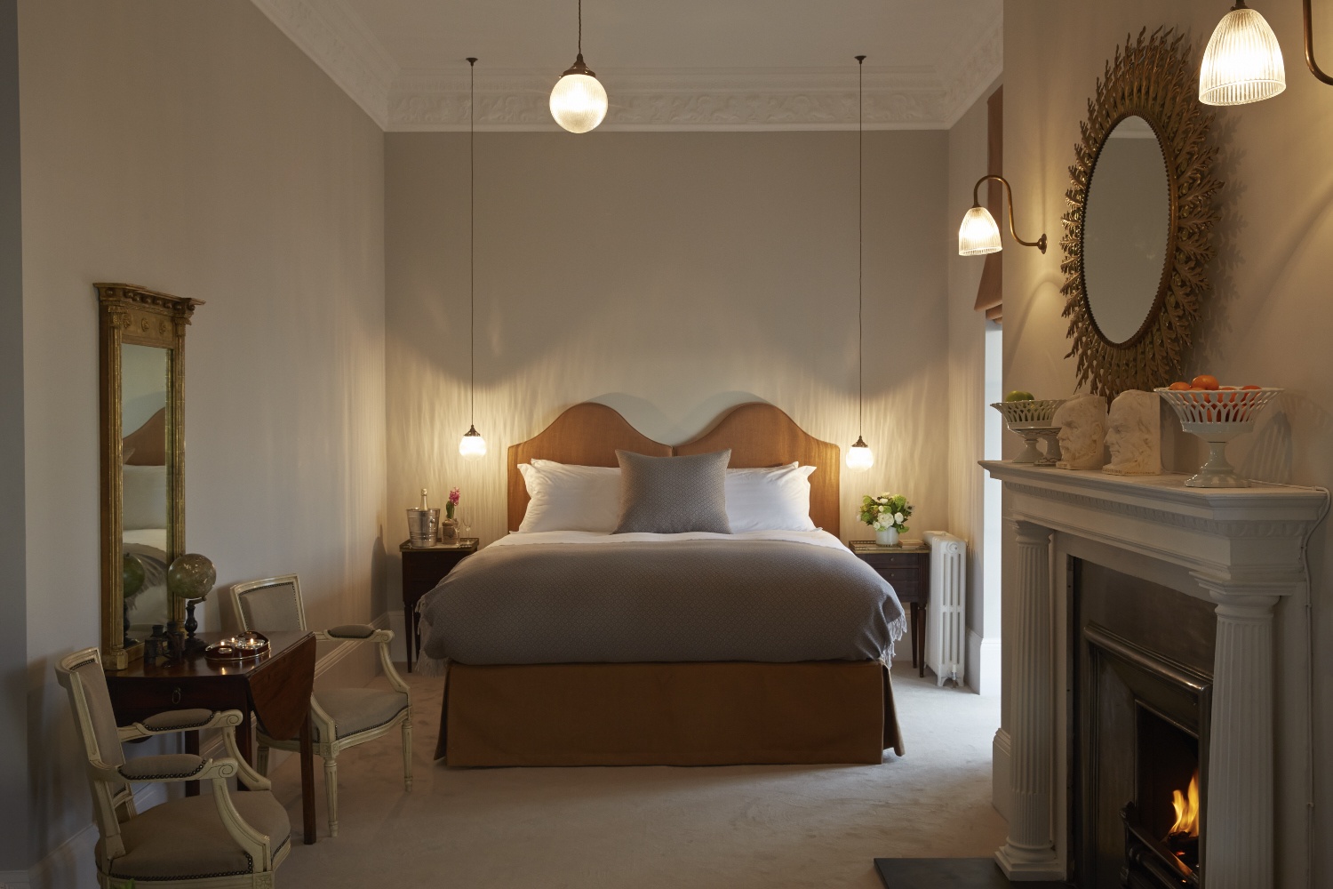 Double bed with white covers, small unit either side and light above hanging down. Wooden headboard and base. On left side mirror with desk and two chairs underneath. On right fireplace with mirror above